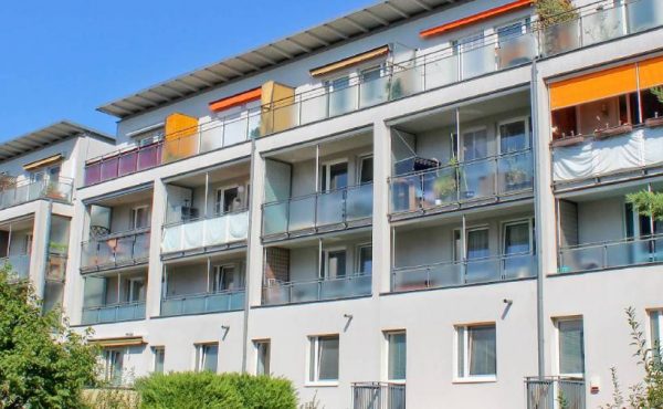 Mehrfamilienhaus mit Balkonen im Grünen bei sonnigem Wetter. Moderne Wohnarchitektur, urbanes Umfeld, blauer Himmel.
