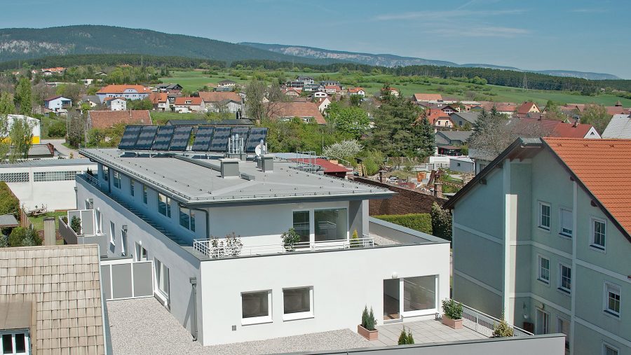 Modernes Gebäude mit Solaranlage auf dem Dach in einem ländlichen Dorf mit grüner Landschaft im Hintergrund.