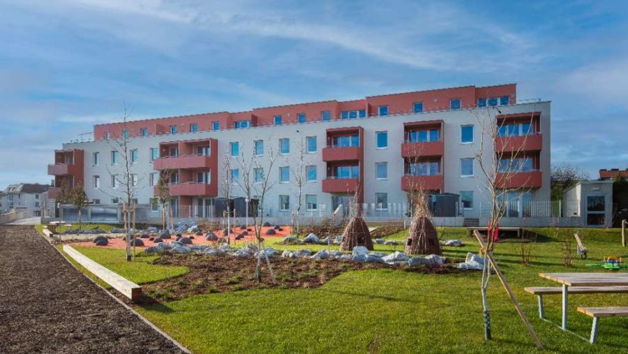 Mehrfamilienhaus mit Balkon und Garten im Vordergrund bei blauem Himmel.