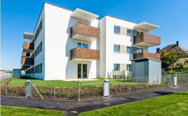 Modernes Mehrfamilienhaus mit weißen Wänden und Balkonen an einem sonnigen Tag, umgeben von grünem Rasen.