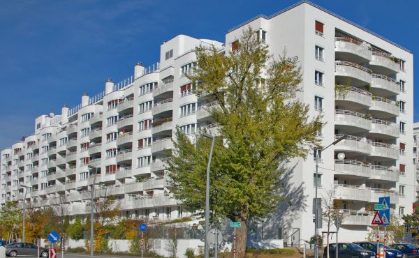 Mehrstöckiges Wohngebäude mit Balkonen und Bäumen, blauer Himmel im Hintergrund.
