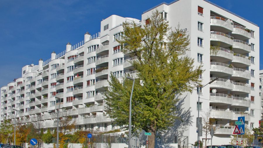 Mehrstöckiges Wohngebäude mit Balkonen und Bäumen, blauer Himmel im Hintergrund.