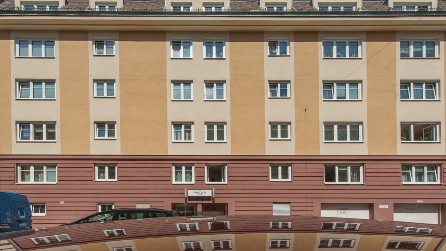 Mehrstöckiges Wohngebäude mit beigen und roten Fassaden, dessen Spiegelung auf einem Autodach sichtbar ist.