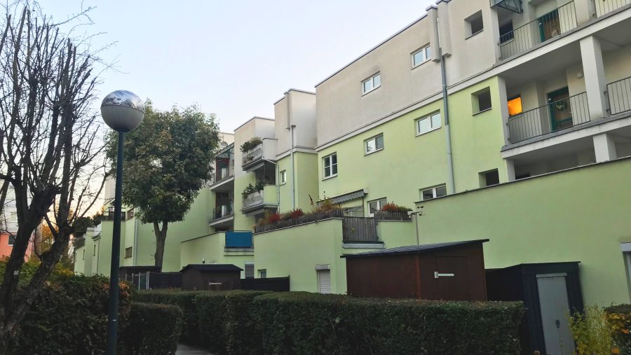 Mehrfamilienhaus mit Balkonen und Garten im Herbst, dazu ein Gehweg und Straßenlaterne.