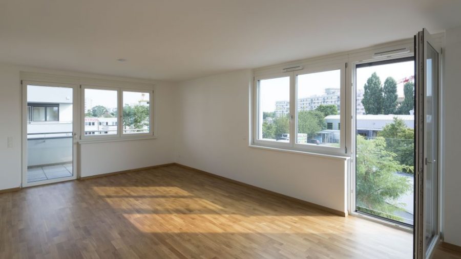 Helles, leeres Zimmer mit Holzboden und großen Fenstern, die auf einen Balkon und eine städtische Aussicht führen.
