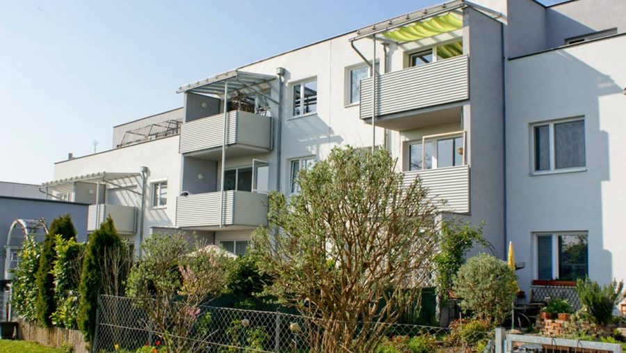 Mehrfamilienhaus mit Balkon und Garten im Vordergrund bei sonnigem Wetter.
