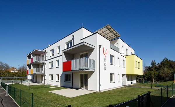 Modernes Wohngebäude mit Balkonen und farbenfrohen Fassaden, umgeben von Grünflächen unter blauem Himmel.