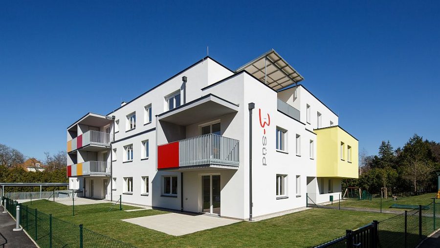 Modernes Wohngebäude mit Balkonen und farbenfrohen Fassaden, umgeben von Grünflächen unter blauem Himmel.