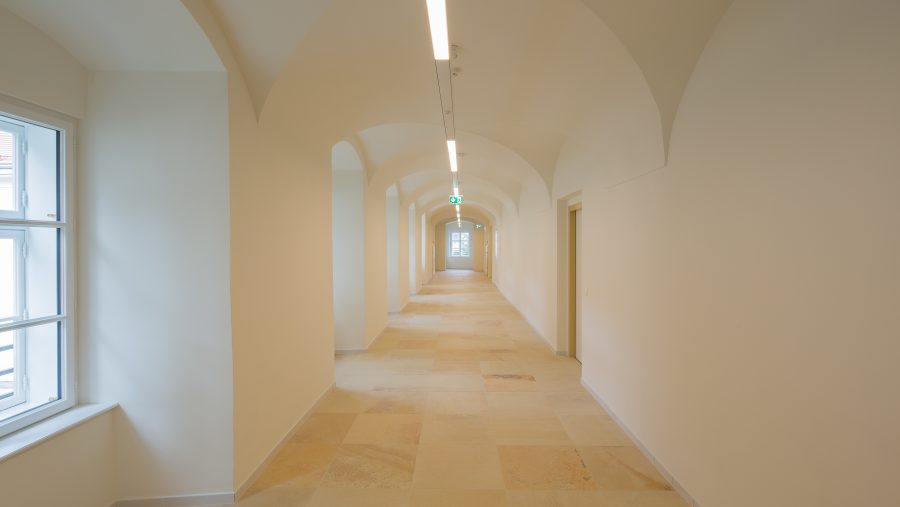 Langer, leerer Flur mit weißem Gewölbe und Fenster an der linken Seite. Helle Beleuchtung und Natursteinboden.