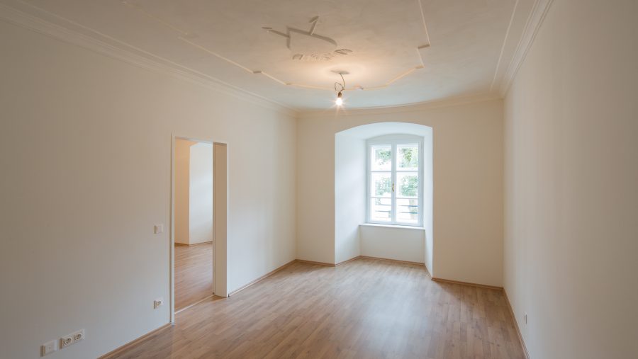 Leerer Raum mit Parkettboden, weißer Wand und Fenster, das Tageslicht hereinlässt.