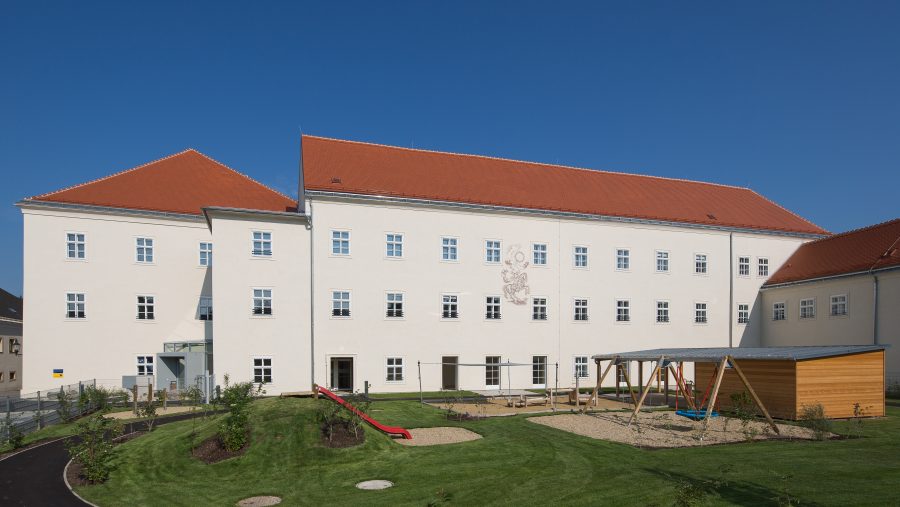 Weißes Gebäude mit rotem Dach und gepflegtem Garten samt Spielplatz unter blauem Himmel.