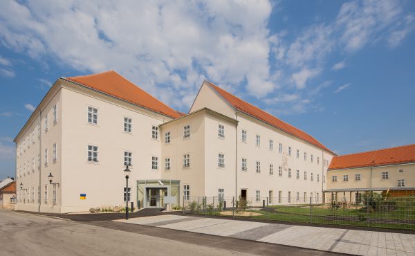 Großes helles Gebäude mit rotem Dach und mehreren Fenstern unter blauem Himmel.