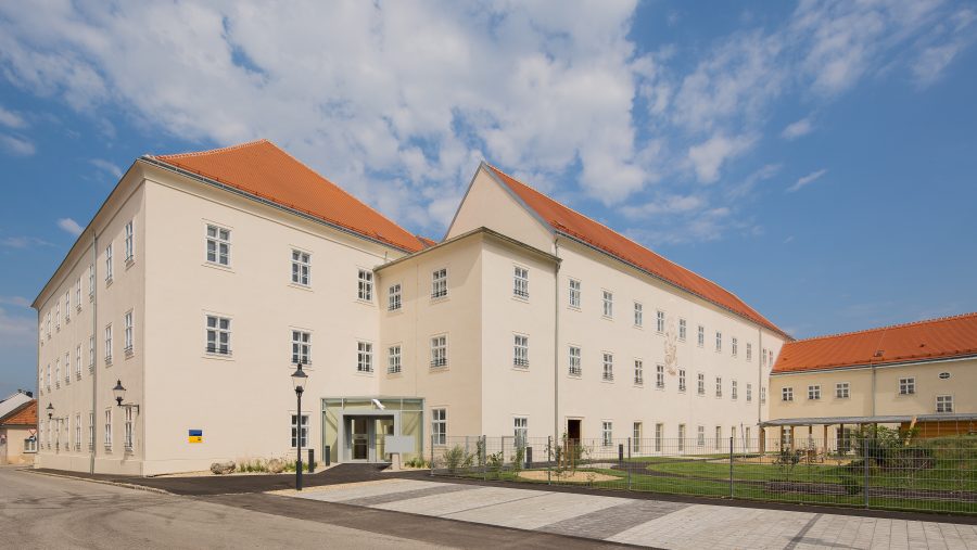 Großes helles Gebäude mit rotem Dach und mehreren Fenstern unter blauem Himmel.
