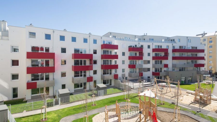 Moderne Wohnanlage mit roten Balkonen und Spielplatz im Innenhof, sonniger Tag.