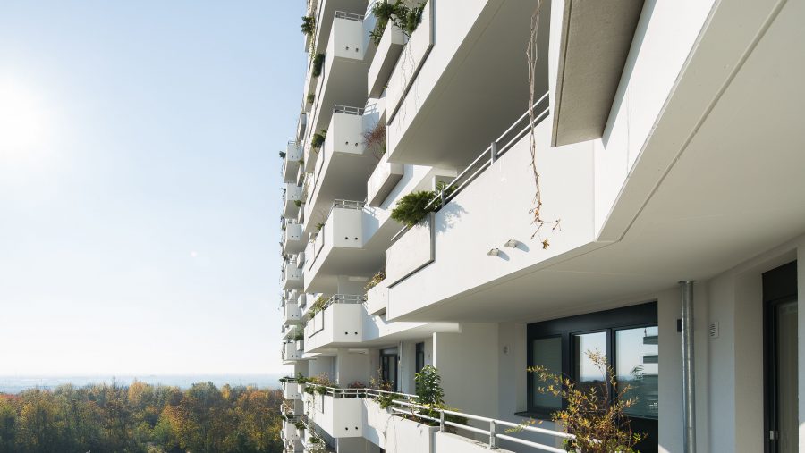Moderne Wohnanlage mit Balkonen, sonniger Tag, Ausblick auf bewaldete Landschaft im Hintergrund.