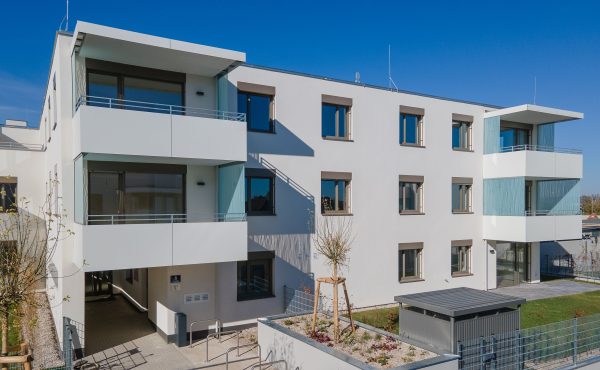 Moderne Wohnanlage mit Balkonen und Garten an sonnigem Tag, blauem Himmel im Hintergrund.