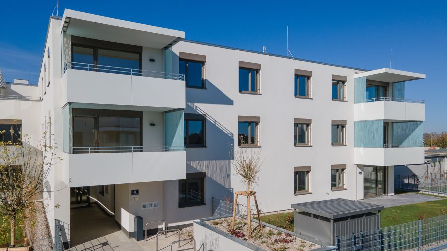 Moderne Wohnanlage mit Balkonen und Garten an sonnigem Tag, blauem Himmel im Hintergrund.