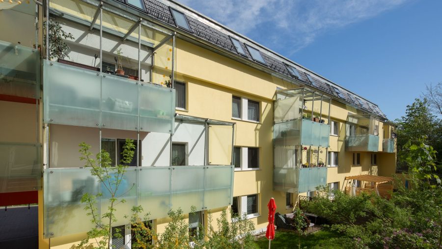 Mehrfamilienhaus mit Balkonen und Solaranlagen, umgeben von grünem Garten unter blauem Himmel.