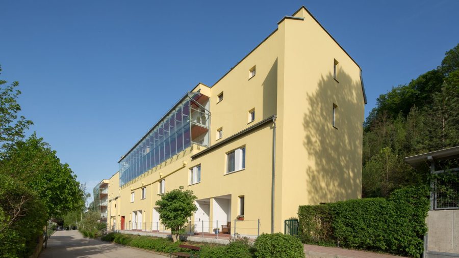 Gelbes Wohngebäude an einem sonnigen Tag, umgeben von Bäumen und blauen Himmel.