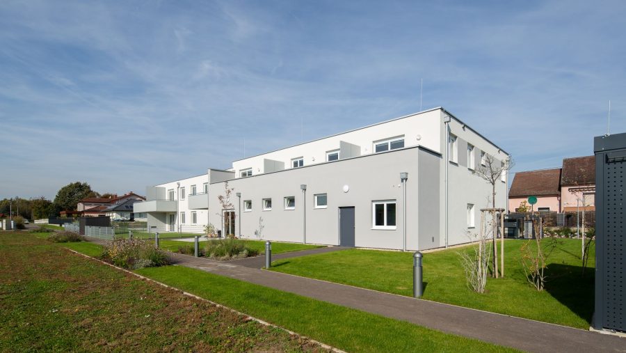 Modernes Wohngebäude bei sonnigem Wetter, mit gepflegtem Rasen und blühenden Pflanzen im Vordergrund.