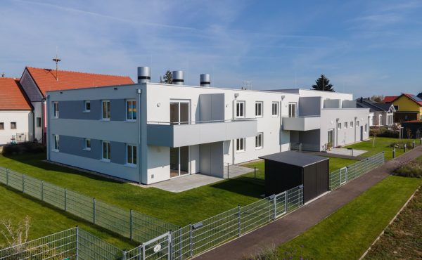 Moderne Wohngebäude mit Garten und Sonnenschein, umgeben von einem Zaun in einer ruhigen Nachbarschaft.