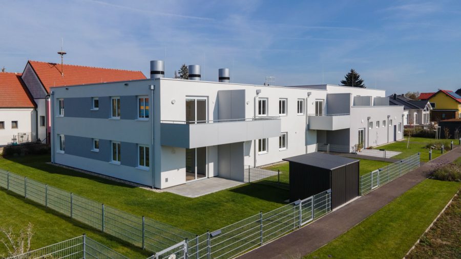 Moderne Wohngebäude mit Garten und Sonnenschein, umgeben von einem Zaun in einer ruhigen Nachbarschaft.