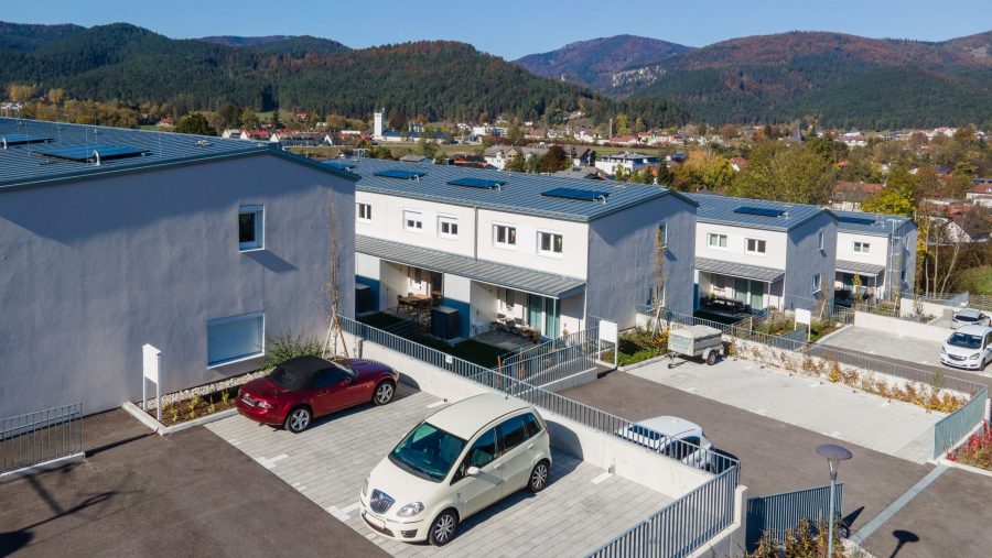 Moderne Reihenhäuser mit Solaranlagen und Parkplatz in einer ländlichen Umgebung mit Bergen im Hintergrund.