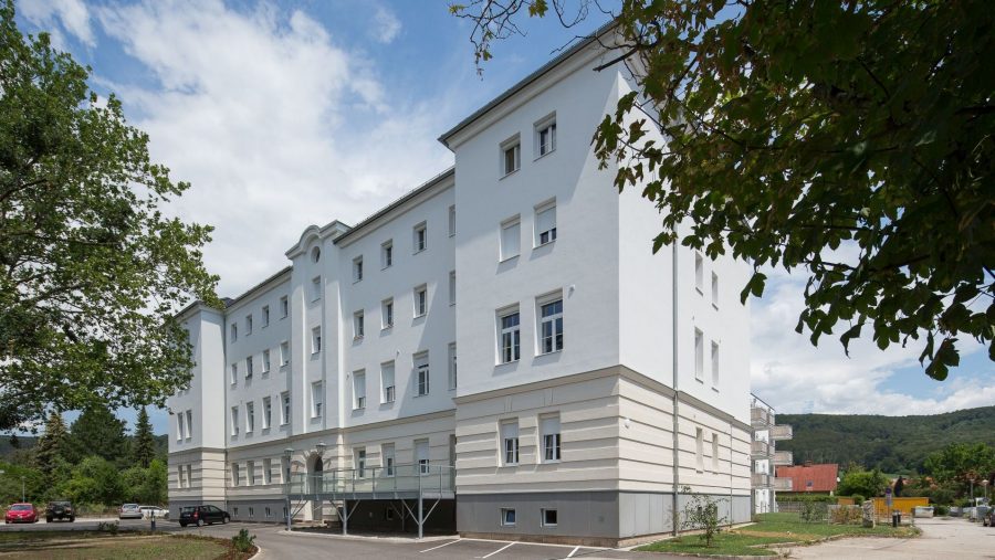 Großes, weißes Gebäude mit mehreren Etagen unter blauem Himmel, umgeben von Bäumen und einem Parkplatz.