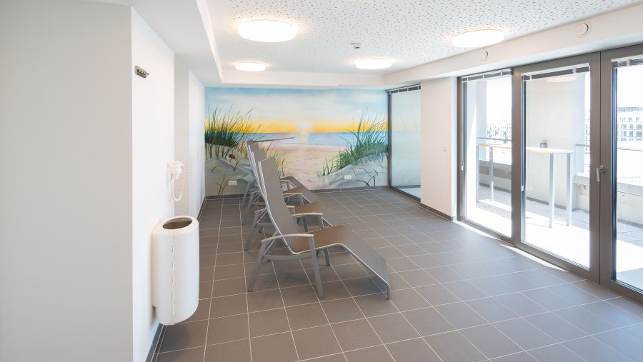 Moderner Ruheraum mit Strandbild an der Wand und Liegestühlen, große Fenster mit Blick ins Freie.