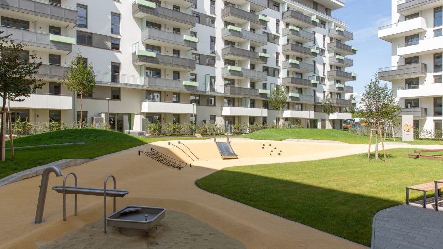 Spielplatz in moderner Wohnanlage mit Sand- und Wasserspielgerät, umgeben von Grünflächen und Mehrfamilienhäusern.