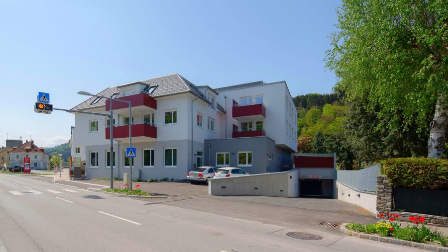 Modernes Wohnhaus an einer Straße mit Ampel und Zebrastreifen, umgeben von Bäumen und einem blauen Himmel.
