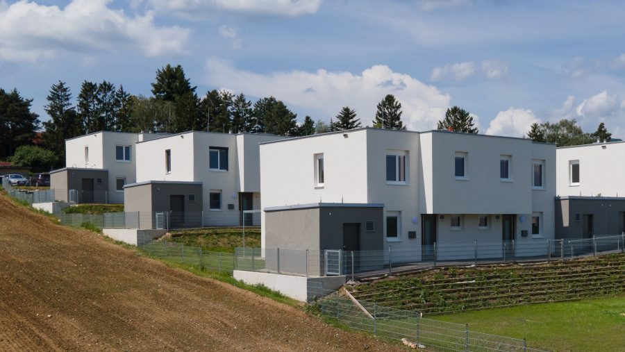 Moderne Reihenhäuser auf einem Hügel, umgeben von grüner Landschaft und Bäumen unter blauem Himmel.