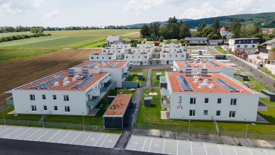 Luftaufnahme einer modernen Wohnanlage mit Solaranlagen auf den Dächern, umgeben von grünen Feldern.