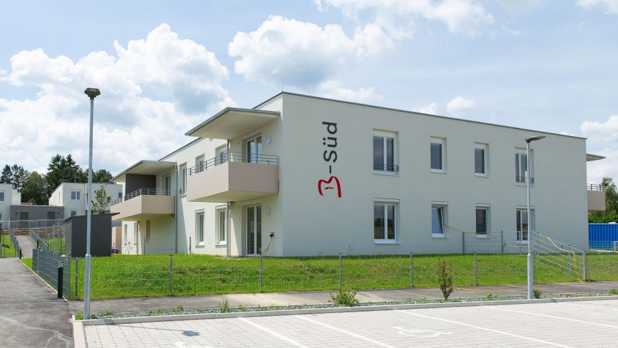 Modernes Gebäude mit Balkon und Parkplatz, umgeben von grünem Rasen unter bewölktem Himmel.
