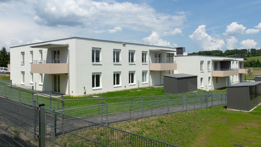 Modernes Gebäude mit mehreren Wohnungen und umliegender grüner Landschaft bei sonnigem Wetter.