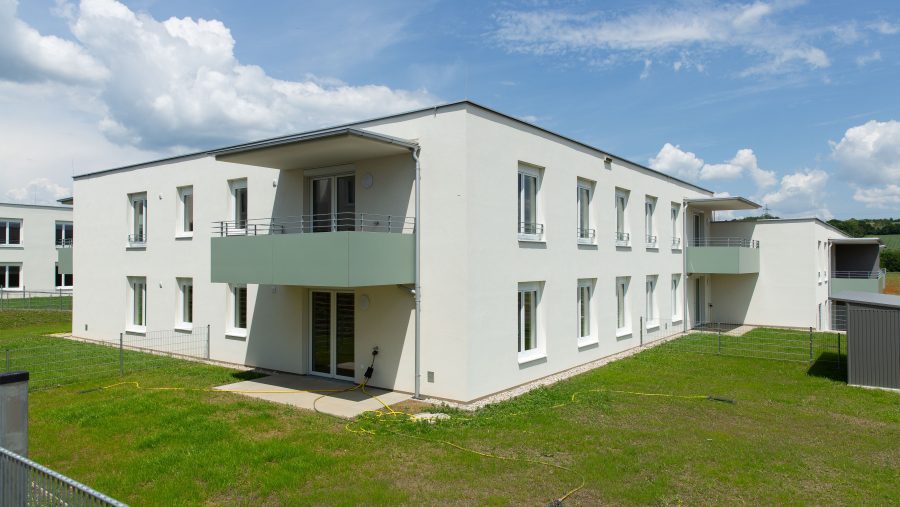 Modernes weißes Gebäude mit Balkon auf einer grünen Wiese, unter blauem Himmel.