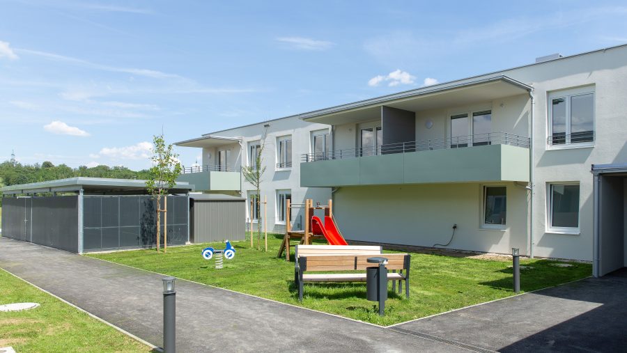 Moderne Wohnanlage mit Balkon, Rasenfläche und Spielplatz bei sonnigem Wetter.