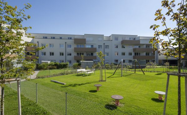 Mehrfamilienhaus mit grünem Innenhof und Spielplatz, sonniger Tag.