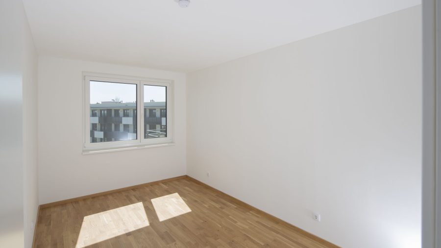 Leeres Zimmer mit Holzboden, weißen Wänden und Fenster mit Blick auf andere Gebäude.