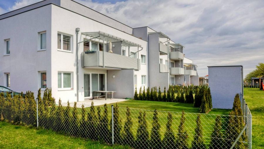Mehrfamilienhaus mit Balkonen und Garten, umgeben von einem Zaun, bei bewölktem Himmel.