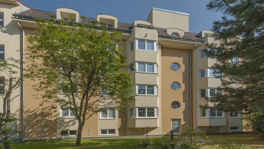 Mehrfamilienhaus mit ockerfarbener Fassade und runden Fenstern, umgeben von Bäumen und Grünfläche bei sonnigem Wetter.