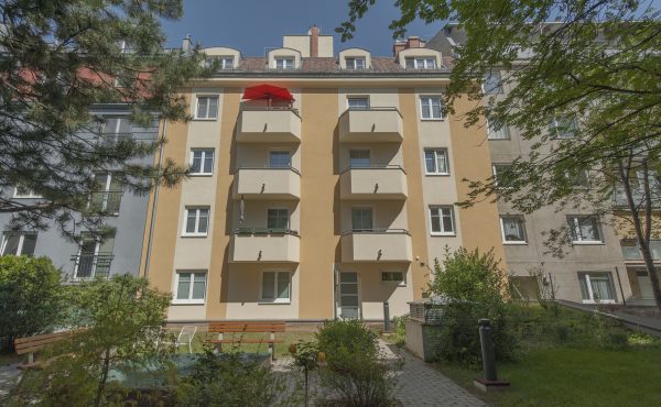 Mehrfamilienhaus mit Balkonen und Garten im Hof, sonniger Tag, moderne Architektur.
