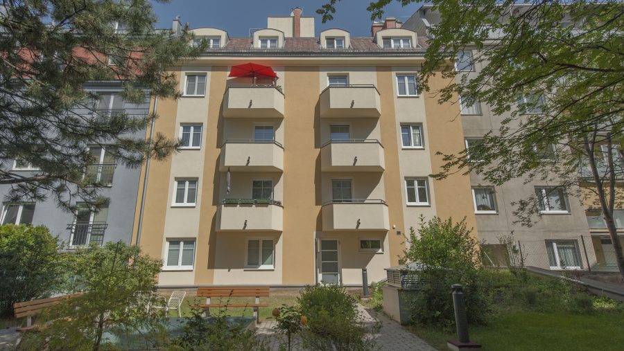 Mehrfamilienhaus mit Balkonen und Garten im Hof, sonniger Tag, moderne Architektur.