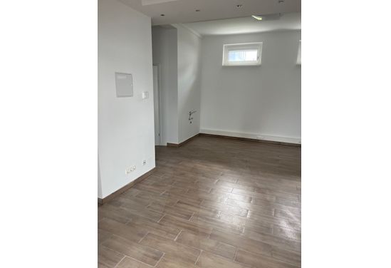 Leerer Raum mit Holzfußboden und weiß gestrichenen Wänden, Tageslicht durch kleine Fenster.