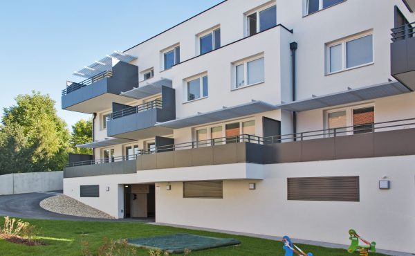 Mehrfamilienhaus mit Balkonen, Spielgeräten im Garten und blauem Himmel im Hintergrund.