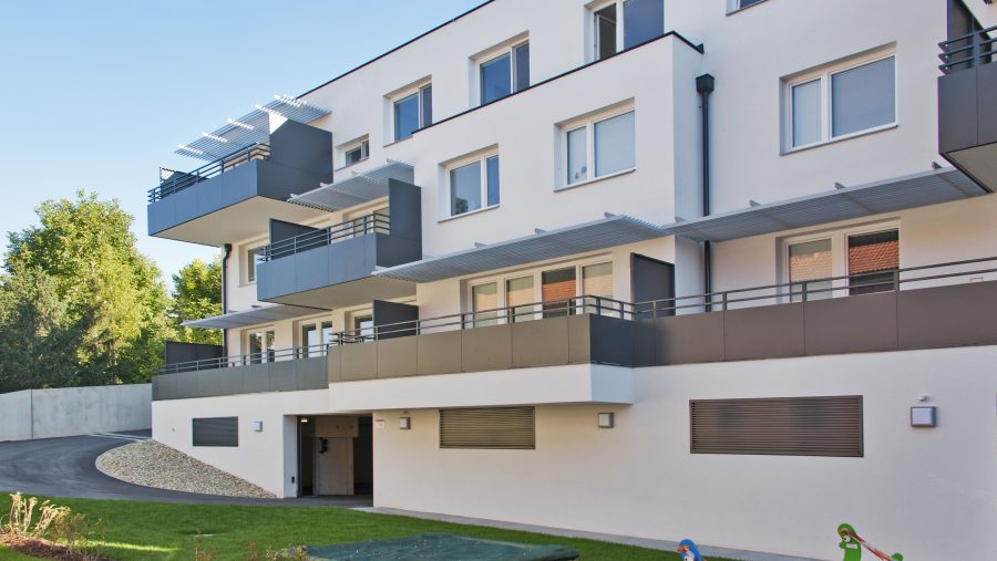 Mehrfamilienhaus mit Balkonen, Spielgeräten im Garten und blauem Himmel im Hintergrund.