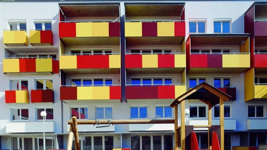 Mehrstöckiges Gebäude mit bunten Balkonen und Spielplatz im Vordergrund unter blauem Himmel.