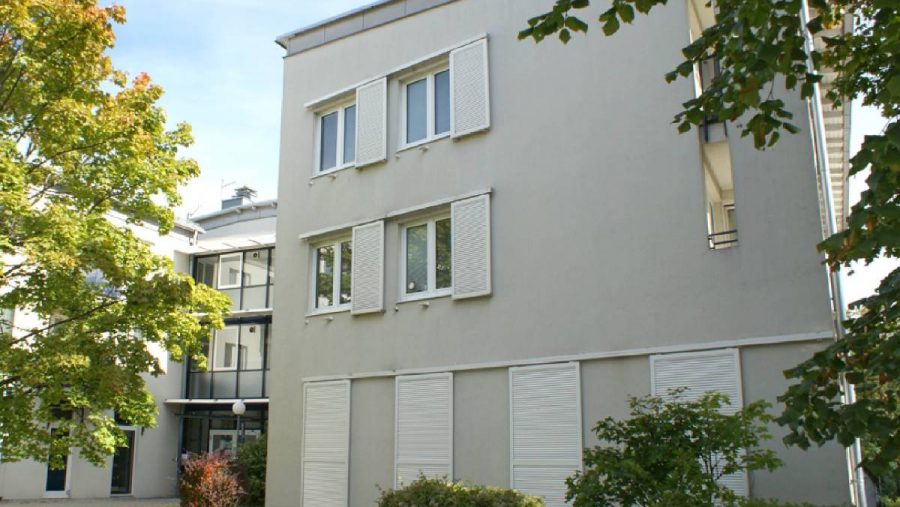 Modernes Gebäude mit geschlossenen Fensterläden, umgeben von grünen Bäumen und Sträuchern im Vordergrund.