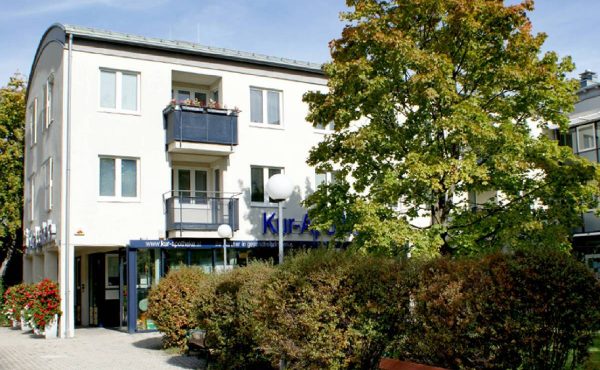 Mehrstöckiges Gebäude mit Balkonen, umgeben von Bäumen und Sträuchern, an einem sonnigen Tag.