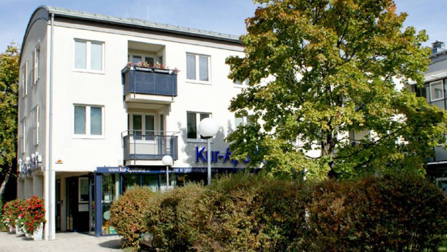 Mehrstöckiges Gebäude mit Balkonen, umgeben von Bäumen und Sträuchern, an einem sonnigen Tag.